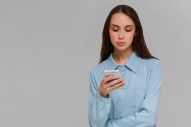 Uzun saçlı, modern cep telefonunu elinde tutan, sosyal ağlarda gezinen, neşeli bir ifadesi olan, gri arka planda izole edilmiş, mesaj alan genç.