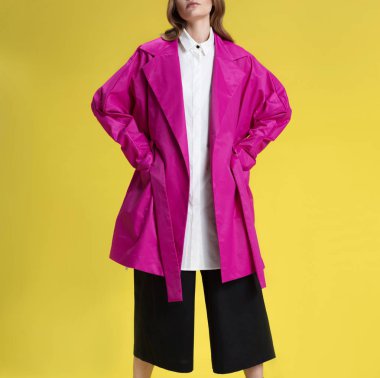 Siyah pantolon, beyaz gömlek ve pembe yağmurluk güzel sarışın kız, sarı arka plan üzerinde poz, stüdyoda çekim