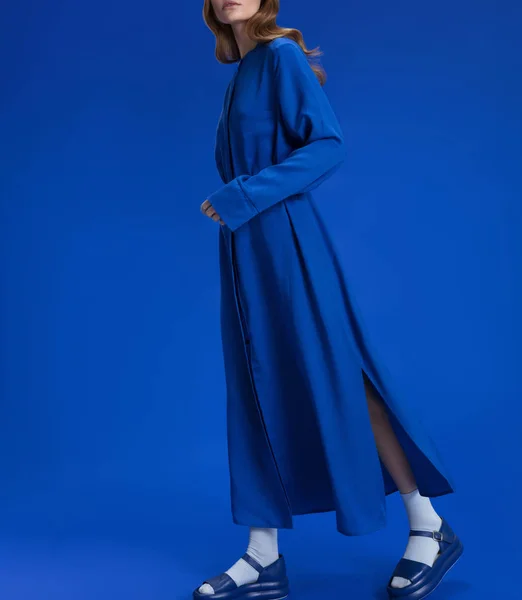 A young girl with blond long hair in a beautiful long blue dress shirt, in blue shoes, white socks on a blue background, studio shot