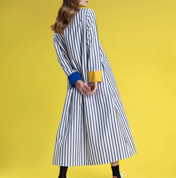 Young girl with long blonde hair in fashionable clothes, black and white with colored inserts long striped shirt dress posing on bright yellow background, studio shot