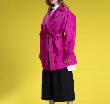 Siyah pantolon, beyaz gömlek ve pembe yağmurluk güzel sarışın kız, sarı arka plan üzerinde poz, stüdyoda çekim