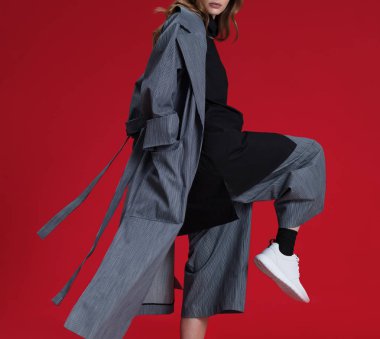 Gri pantolon ve siyah gömlek ve kar yağmurluk, beyaz spor ayakkabı, kırmızı arka plan üzerinde poz güzel sarışın kız, stüdyoda çekim