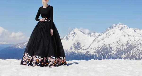 Beautiful photo of a young girl model in a black net dress with embroidery floral print, which stands on top of a snow-covered mountain overlooking the beautiful mountains