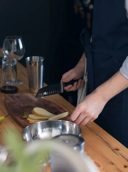 Male bartender in a blue apron in the kitchen, cutting fruits on a wooden cutting board