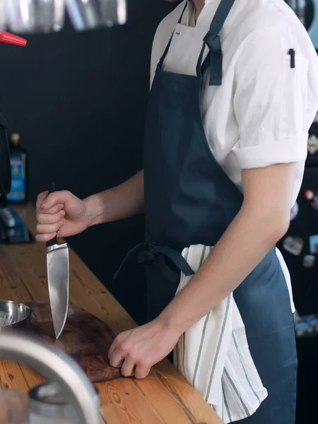 A male cook in a blue apron in the kitchen, stuck a knife into a wooden chopping board