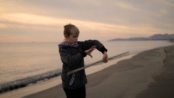 日落时, 男孩站在海滩上, 转动手中的棍子. — 图库视频影像