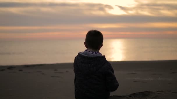 Ten chlapec sedí na pláži při západu slunce a hodí kámen do moře. — Stock video