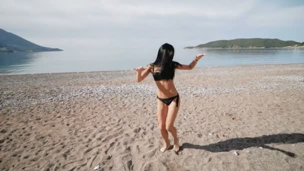 Сексуальна дівчина в чорному бікіні купальнику з красивою спортивною фігурою танцює на березі моря — стокове відео