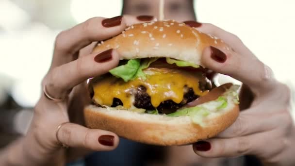 A young girl has a big appetizing cheeseburger. Holds it in the hands of a close-up. A woman bites him and gets great pleasure. Close-up. Slowmotion 60fps — Stock Footage Young Girl Has Big Appetizing Cheeseburger Holds Hands Close Woman Royalty Free Stock Video
