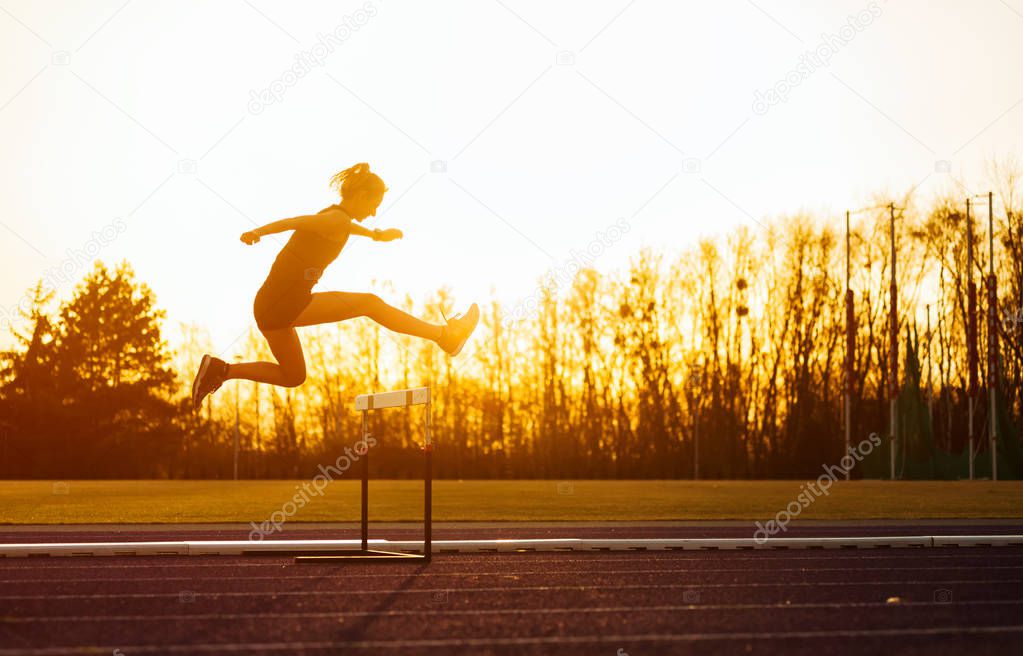 Athletic woman running on stadium track and jumping above hurdle at sunset