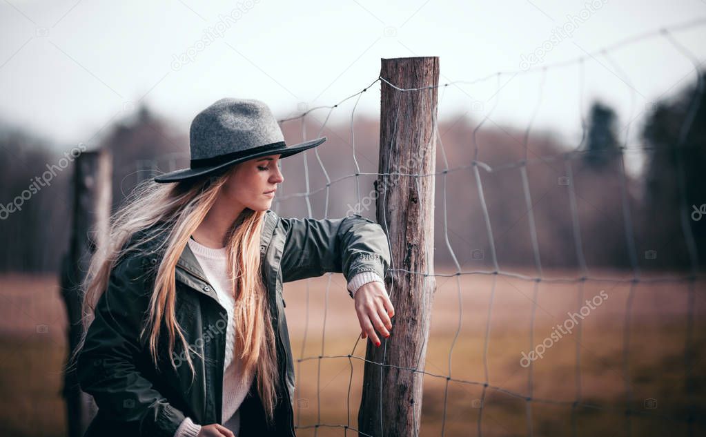 Young woman in stylish hat posing near rural fence