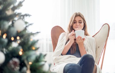 Genç, rahat bir kadın Noel ağacının yanında dinleniyor ve sıcak çay ya da kahve içiyor.