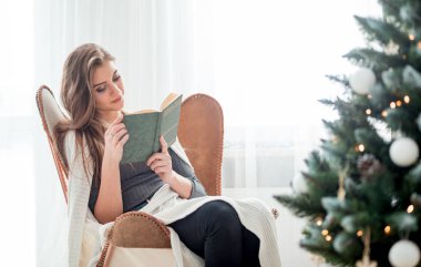 Genç ve güzel bir kadın Noel ağacının yanında oturmuş kitap okuyor.