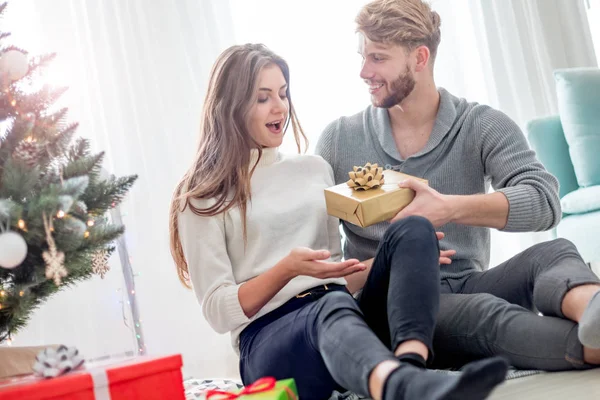 幸せなカップルはクリスマスの木の隣の床に座って自分自身にクリスマスプレゼントを与えています — ストック写真