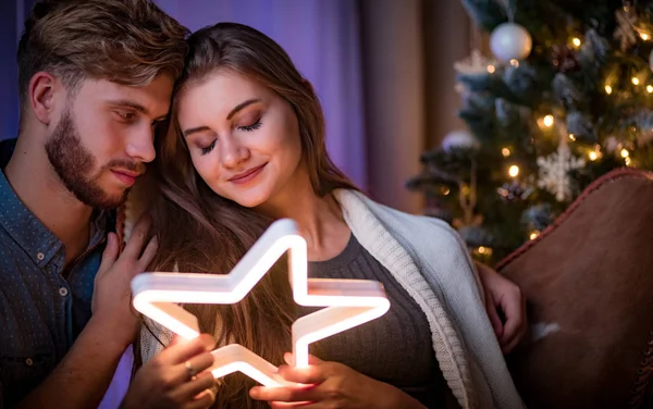 欢快的夫妇坐在圣诞树旁边 在圣诞前夜的黑暗舒适的房间里抱着发光的Led星星 — 图库照片