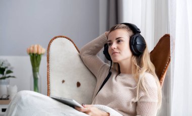Woman at home wearing headphones while using digital tablet, listening to audiobooks or music clipart