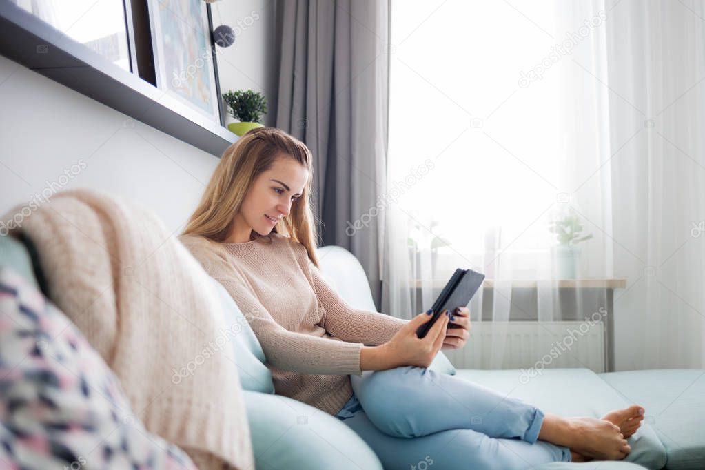 Happy woman reading ebook on digital reader while lying on comfortable sofa at home, leisure time