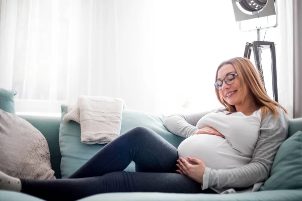 Mladá těhotná žena sedí na gauči v obývacím pokoji — Stock fotografie