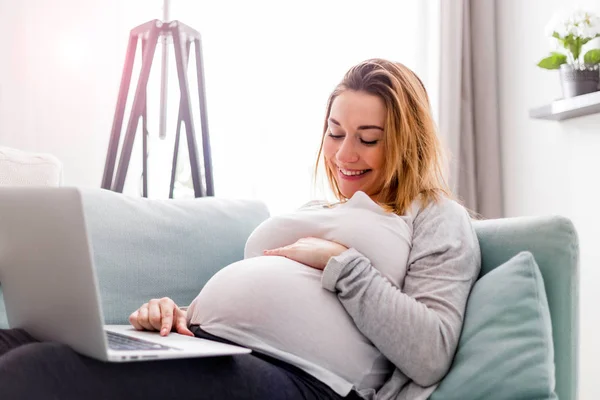 Mosolygó terhes nő használ laptop, ül a kanapén otthon — Stock Fotó