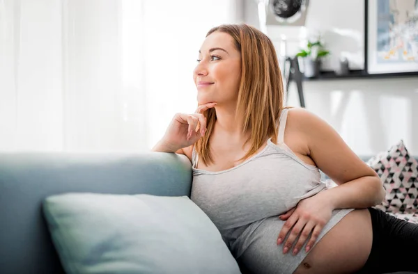 Улыбающаяся беременная женщина сидит на диване и смотрит в окно, ожидая ребенка концепции — стоковое фото