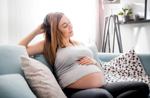 Entspannte Schwangere sitzt zu Hause auf Sofa und hört Musik über Kopfhörer — Stockfoto