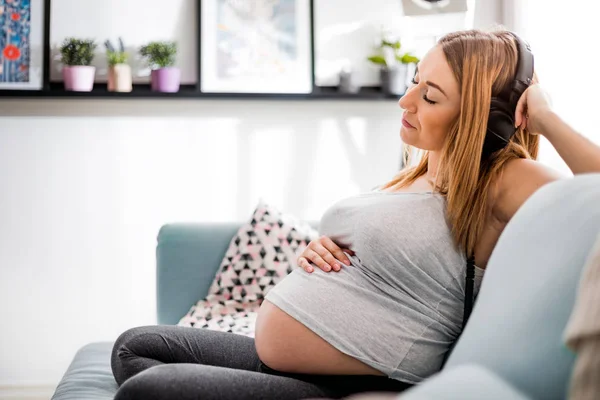 Uvolněná těhotná žena sedí na pohovce doma a poslouchá hudbu ve sluchátkách — Stock fotografie