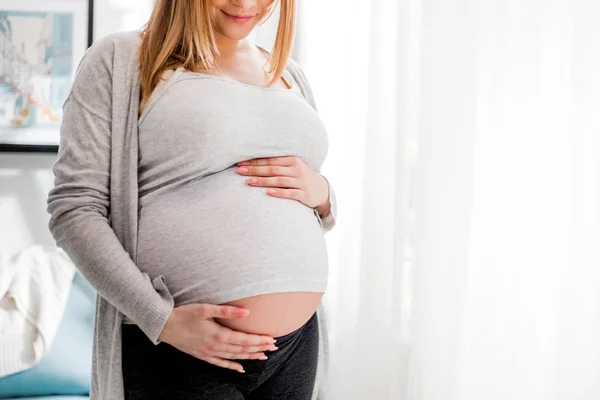 Těhotná žena, která se dotýká jejího břicha, stála u okna doma — Stock fotografie