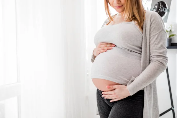 Terhes nő megérintette hasa állt az ablak otthon — Stock Fotó