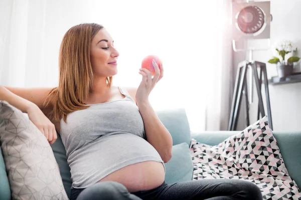 Terhes nő étkezési alma és pihenő ül a kanapén otthon — Stock Fotó