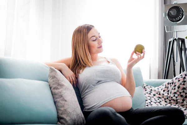 Výživa a strava v těhotenství, těhotné ženy s ovocem, které sedí na pohovce doma — Stock fotografie