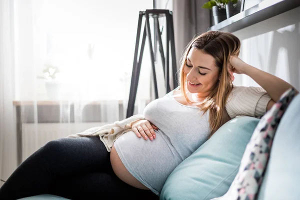 Schöne schwangere junge Frau berührt ihren Bauch sitzt auf dem Sofa zu Hause — Stockfoto