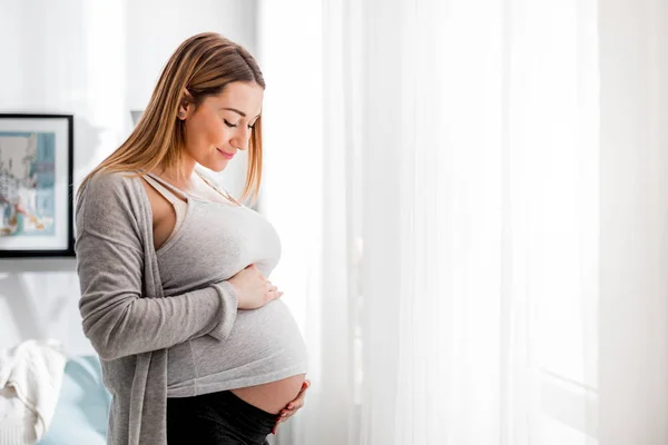 Красивая беременная женщина трогает живот, стоя у окна дома. — стоковое фото