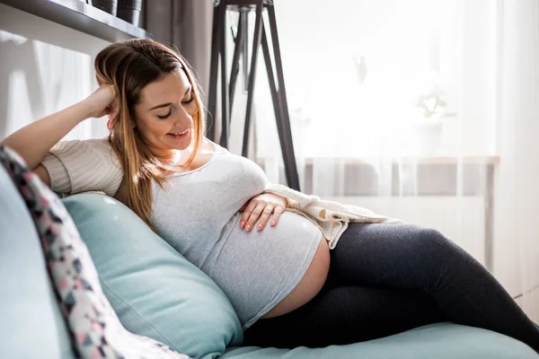 Krásná těhotná mladá žena, která se dotýká břicha, seděla doma na pohovce — Stock fotografie