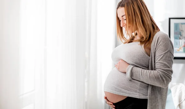 Красивая беременная женщина трогает живот, стоя у окна дома. — стоковое фото