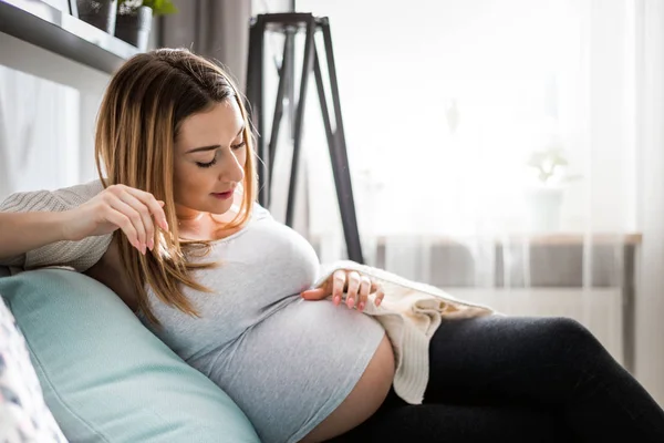 Schöne schwangere junge Frau berührt ihren Bauch sitzt auf dem Sofa zu Hause — Stockfoto