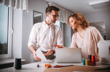 Moda tasarım stüdyosunda dizüstü bilgisayar ve dikiş makinesi kullanarak proje çalışmaları sırasında profesyonel terziler