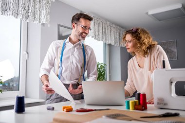 Moda tasarım stüdyosunda dizüstü bilgisayar ve dikiş makinesi kullanarak proje çalışmaları sırasında profesyonel terziler