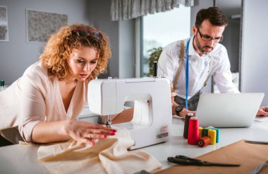 Moda tasarım stüdyosunda dizüstü bilgisayar ve dikiş makinesi kullanarak proje çalışmaları sırasında profesyonel terziler