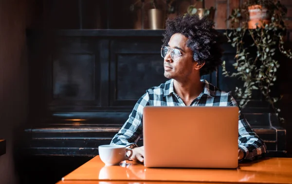 Hombre africano en gafas usando portátil mientras está sentado en el restaurante, concepto de los jóvenes que trabajan — Foto de Stock