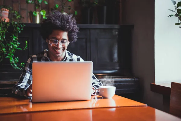 Hombre africano en gafas usando portátil mientras está sentado en el restaurante, concepto de los jóvenes que trabajan — Foto de Stock