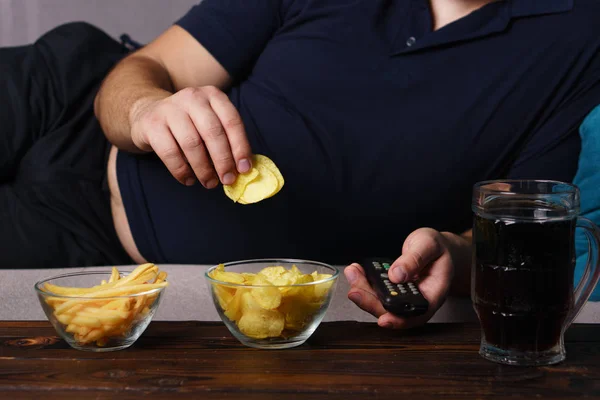 Hombre con sobrepeso con control remoto de televisión, comida chatarra y cerveza — Foto de Stock