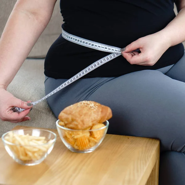 Stress, problemi alimentari, sovralimentazione compulsiva, aumento di peso. donna in sovrappeso con nastro adesivo in vita e cibo spazzatura — Foto Stock