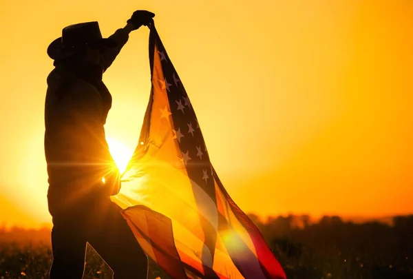 男性ウエスタン風光明媚な日没の眺めの中に大規模な米国旗を振って独立記念日を祝っている身に着けています カウボーイ ハットでアメリカ合衆国のパトリオット — ストック写真