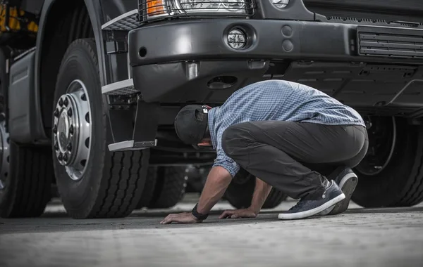 壊れたトラック 代の白人のトラック運転手の問題彼の車の下で損害を探しています 自動車および輸送業界テーマ — ストック写真