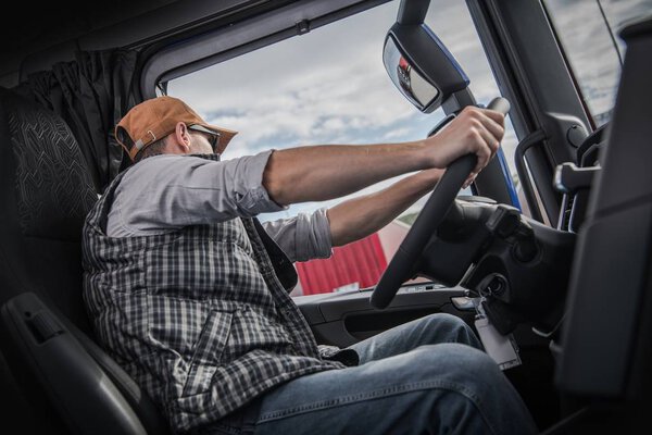 Caucasian Trucker in His 30s Behind Modern Semi Truck Wheel. Transportation Industry.