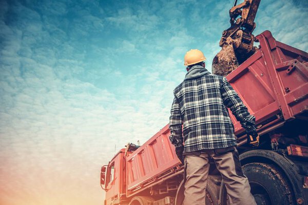 Ground Works Operation. Construction Worker in Front of Dump Truck Loaded by Excavator. Heavy Duty Machinery. Soil Removing.