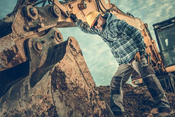 Construction Industry. Heavy Equipment Operator Job. Excavator and Caucasian Worker in Hard Hat. Men Looking Inside Excavator Bucket.