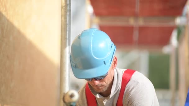 Blanke bouwvakker in blauwe harde hoed op een steiger die zijn werk doet. — Stockvideo