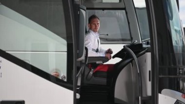 Beyaz tenli otobüs şoförü güneş gözlükleriyle kulübede oturuyor. Aracın girişine bakıyor ve dışarıda bekleyen yolcuyla konuşuyor.. 