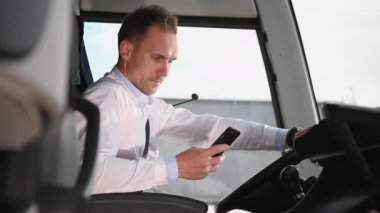 Erkek koç molada. Park halindeki otobüsün kabininde oturuyor. Cep telefonunu kontrol ediyor.. 
